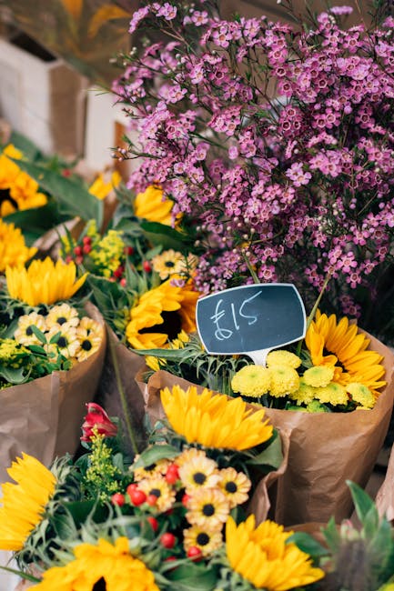 A vibrant display of fresh floral arrangements featuring bright yellow sunflowers and small daisy-like flowers wrapped in brown paper, set outdoors with a purple-pink flowering shrub in the background