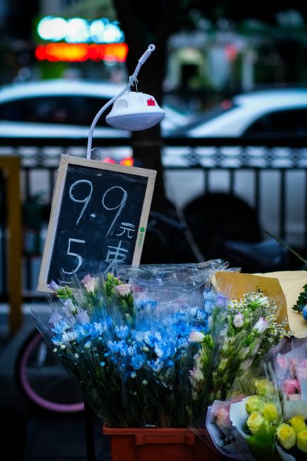 A display of fresh flower bouquets arranged in a plastic container at an outdoor flower stall, featuring blue and pink blooms with green foliage, wrapped in clear plastic. A black chalkboard sign with