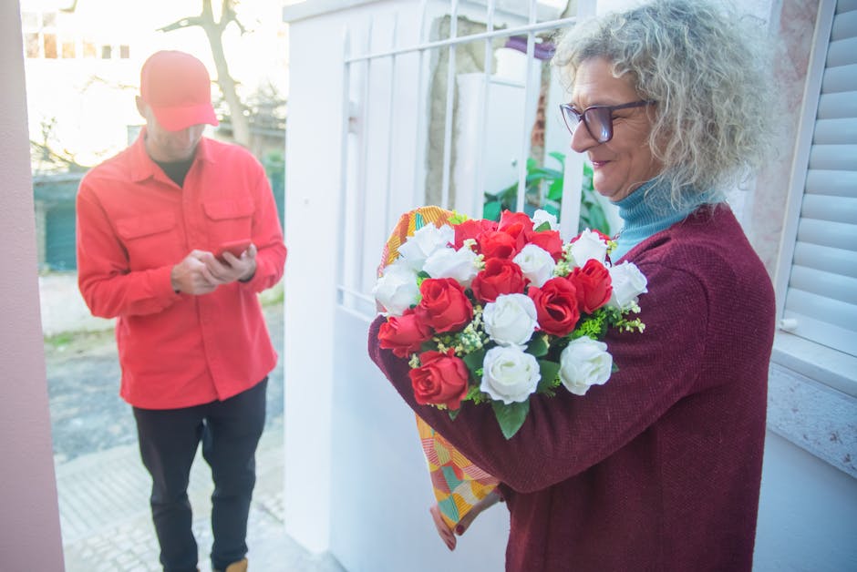 A woman with curly gray hair and glasses, dressed in a burgundy cardigan and blue turtleneck, is holding a vibrant bouquet of fresh red and white roses wrapped in colorful patterned paper outside a do
